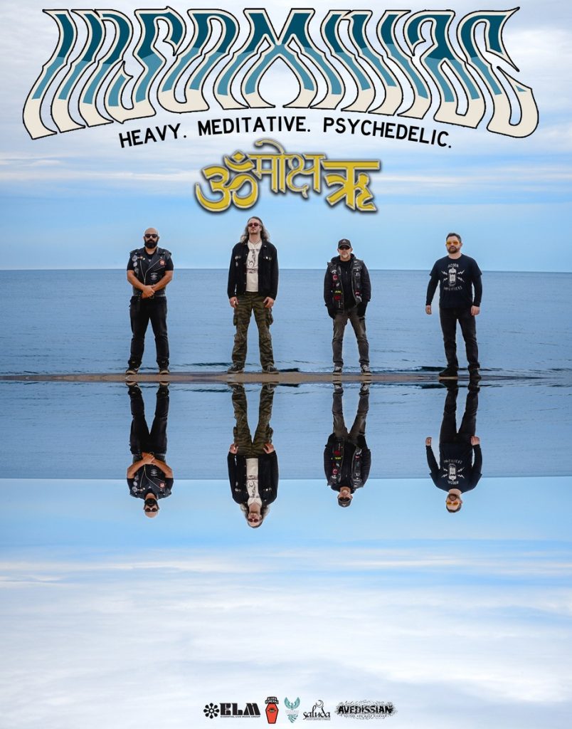 Four musicians stand on a beach, reflecting in the water, with a cloudy sky above.