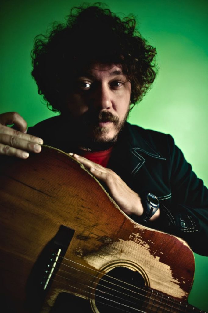 A man with curly hair holds a worn guitar, gazing thoughtfully at the camera against a green background.