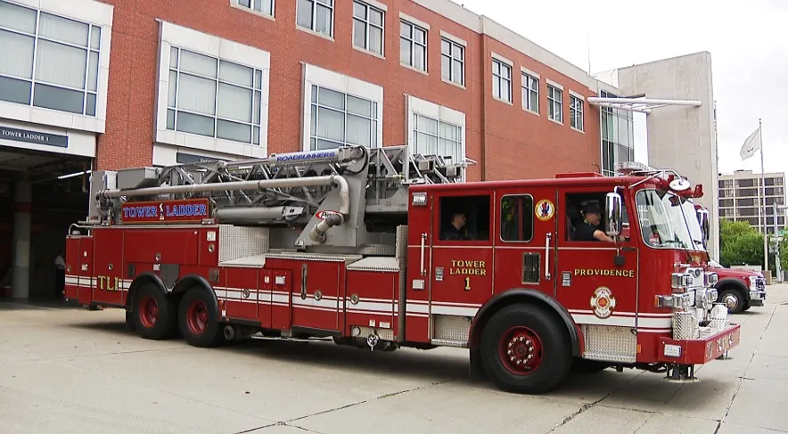 A red fire truck with a ladder is parked outside a fire station, with firefighters visible inside.