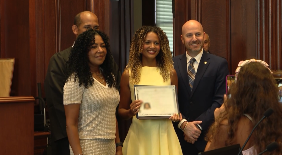 A woman in a yellow dress holds a certificate, surrounded by three smiling individuals in a formal setting.