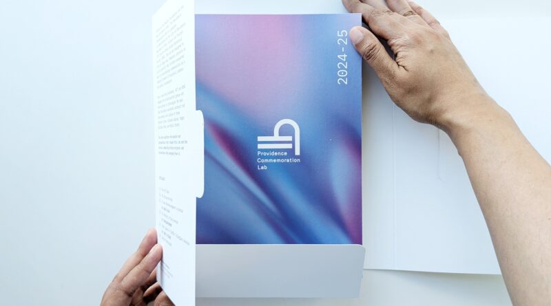 A hand opens a folder revealing a colorful document titled "2024-25 Providence Commemoration Lab.