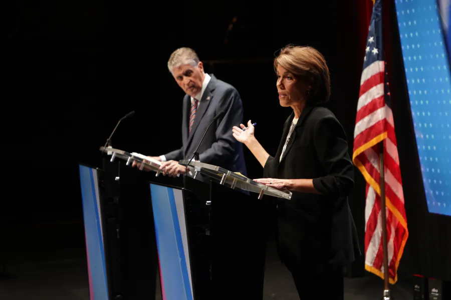 Two candidates debate at podiums, one gesturing while the other listens, with an American flag in the background.