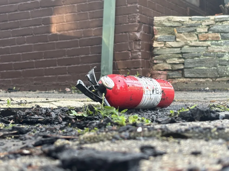 A red fire extinguisher lies on the ground near charred debris and green grass.