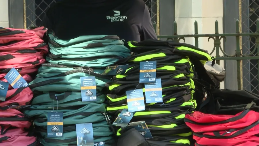 Stacks of colorful backpacks with tags, displayed outdoors near a person wearing a Beacon Bank shirt.