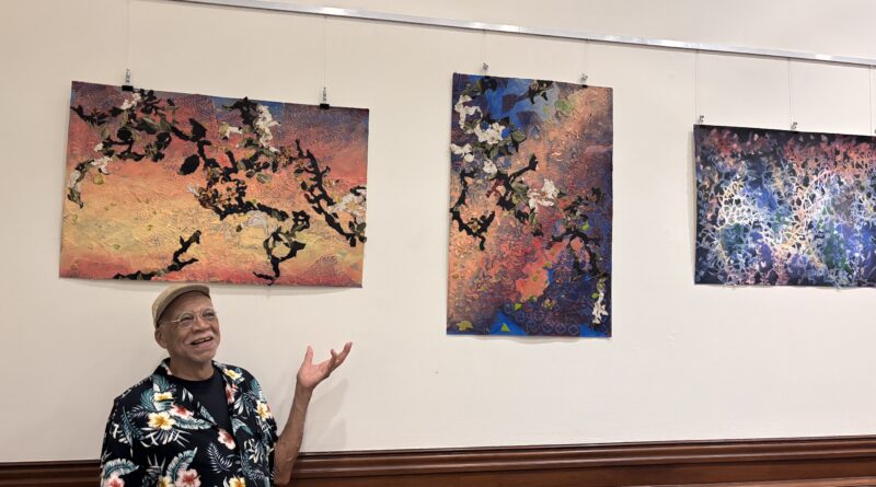 An older man in a floral shirt gestures towards three colorful paintings on the wall.