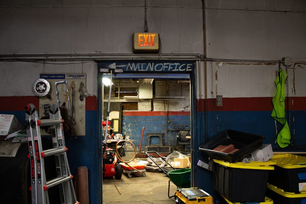 A cluttered workshop with an exit sign, ladder, and various tools visible. Main office door is open.