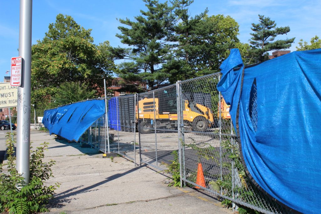 Construction site with blue tarps, orange machinery, and a chain-link fence under a clear blue sky.