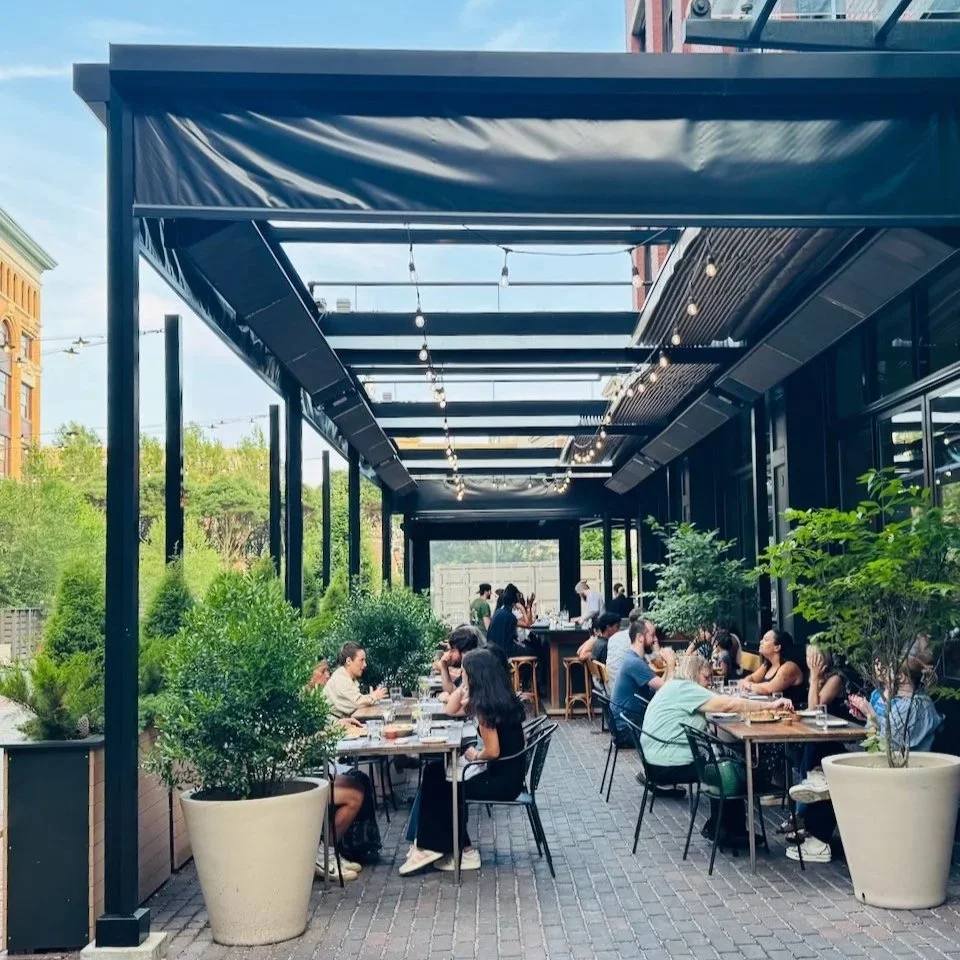 Patio dining scene with people at tables, greenery, and string lights overhead.