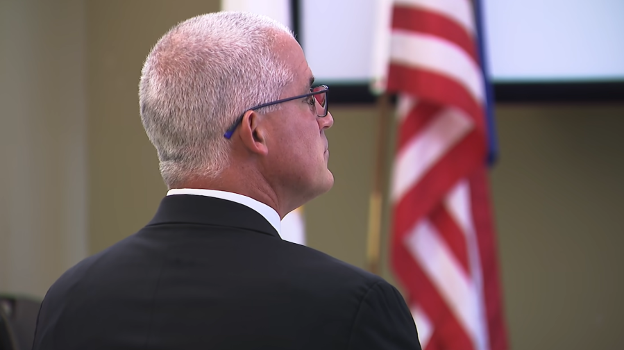 A man in a suit listens attentively, facing away from the camera, with an American flag in the background.