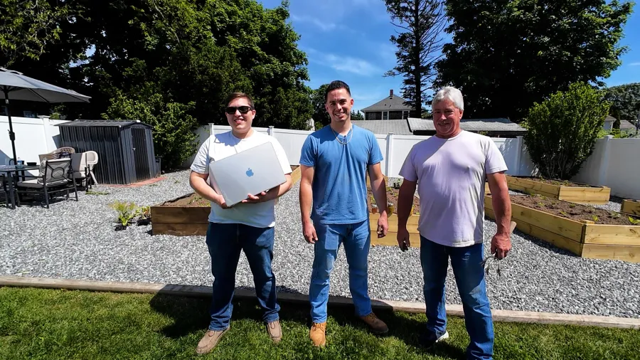 Three men stand in a garden, one holding a laptop, with raised garden beds and a patio in the background.