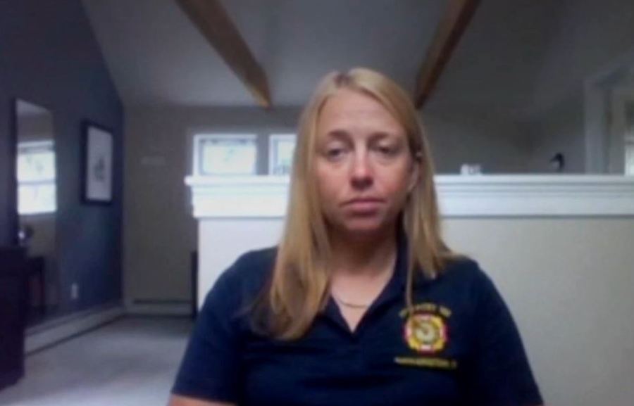 A woman with long hair sits in a room, looking directly at the camera, wearing a dark shirt with a logo.