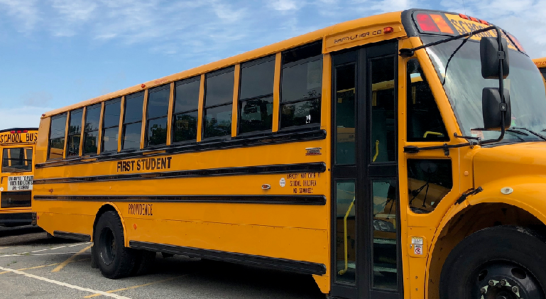 Yellow school bus parked, labeled "First Student," with clear windows and a blue sky in the background.