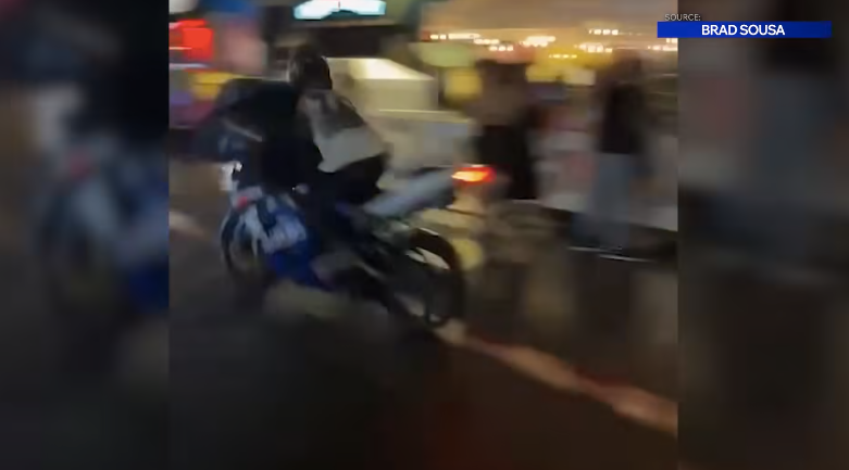 A person rides a motorcycle through a busy street at night, with pedestrians nearby.