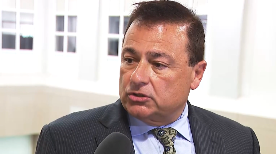 A man in a suit speaks into a microphone, with a bright interior background.