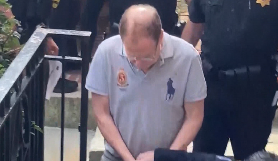 A man in a gray polo shirt is escorted by police officers, looking down as he walks.