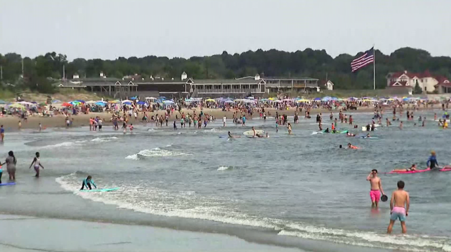 Crowded beach scene with people swimming, surfing, and umbrellas. An American flag waves in the background.