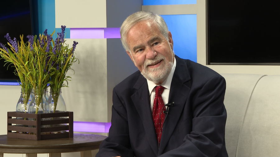 A smiling older man in a suit sits on a couch, with a vase of flowers nearby.