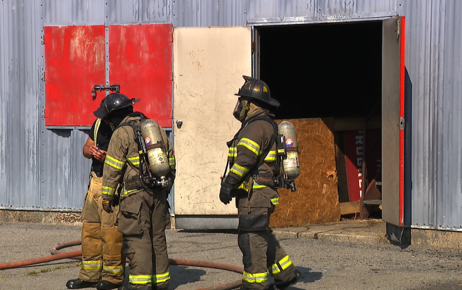 Two firefighters in gear discuss equipment outside a building with open doors. A hose lies on the ground.
