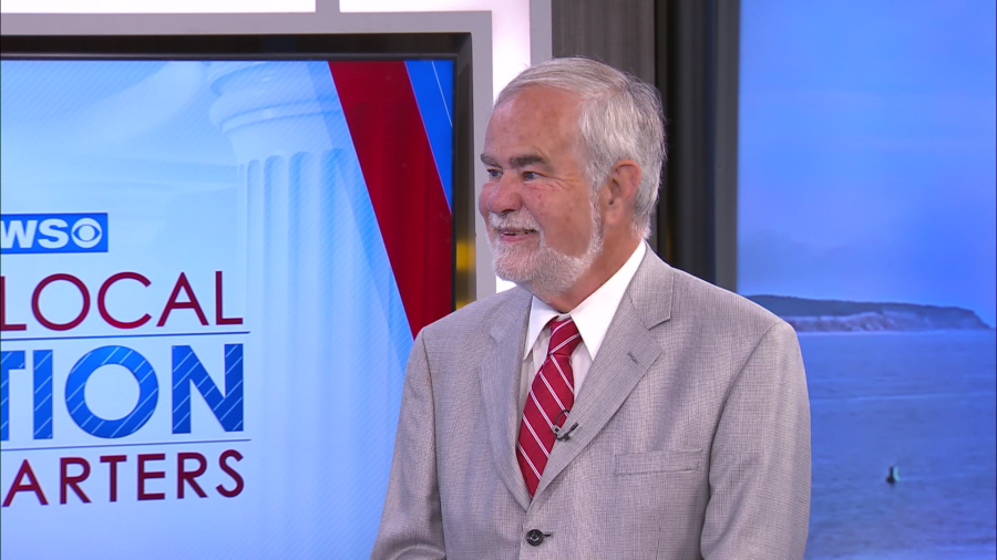 A man in a light gray suit and red tie smiles during a news segment. Background features a coastal view.