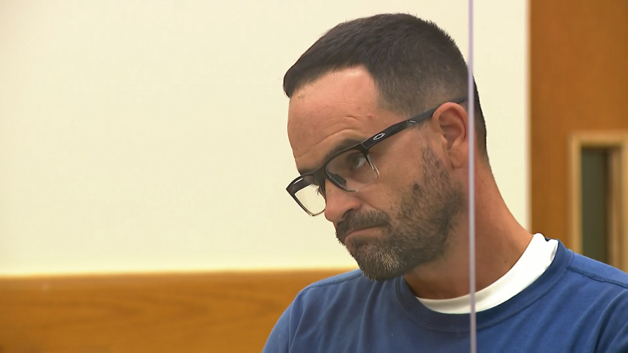 A man with glasses looks down, appearing contemplative in a courtroom setting.