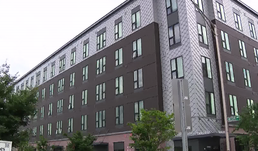 Modern multi-story building with large windows, surrounded by trees and street signs.