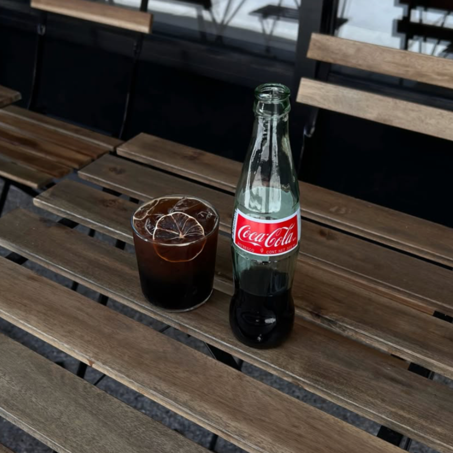 A glass of iced coffee with lemon slices and a bottle of Coca-Cola on a wooden table.