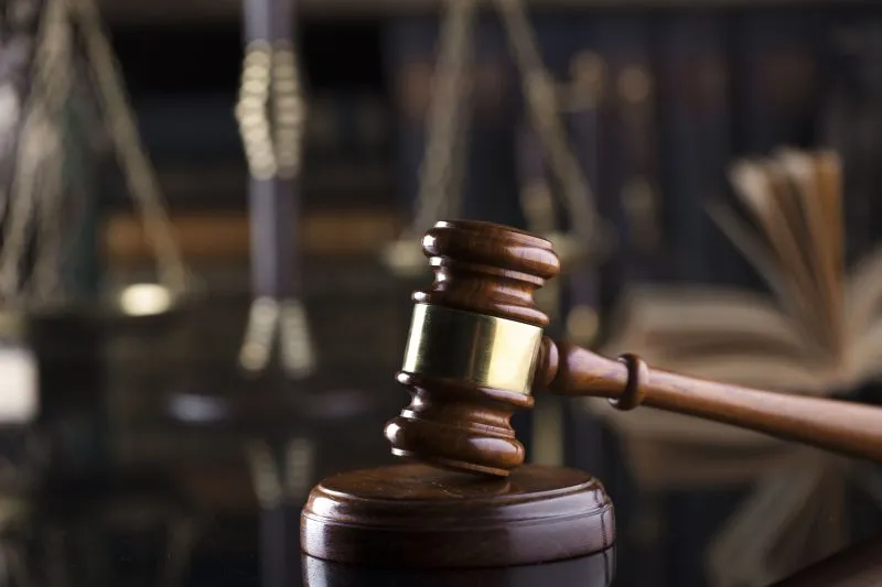 A wooden gavel rests on a reflective surface, with scales of justice and books in the background.