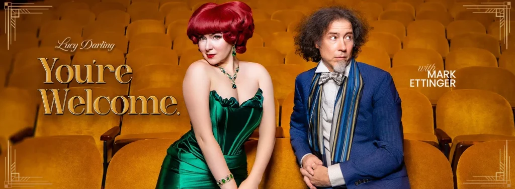 Lucy Darling poses in a green dress beside Mark Ettinger, both seated in a theater with empty orange seats.