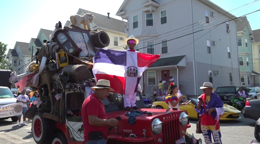 A person in a white hat holds a Dominican flag on a decorated vehicle during a parade. Spectators gather nearby.