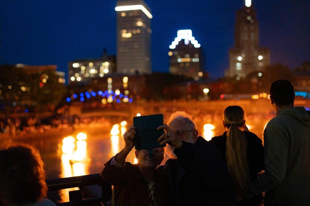 Two people take a selfie by a river at night, with city lights and flames reflecting in the water.