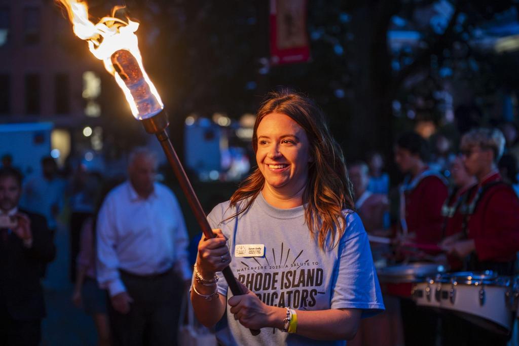 A woman in a blue shirt smiles while holding a flaming torch at an outdoor event.