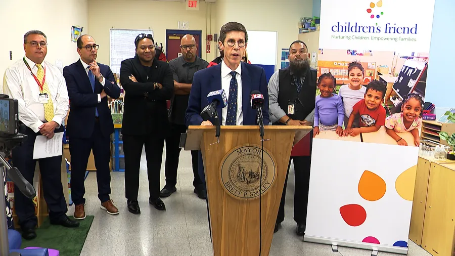 A man speaks at a podium with microphones, surrounded by several people in a children's center.