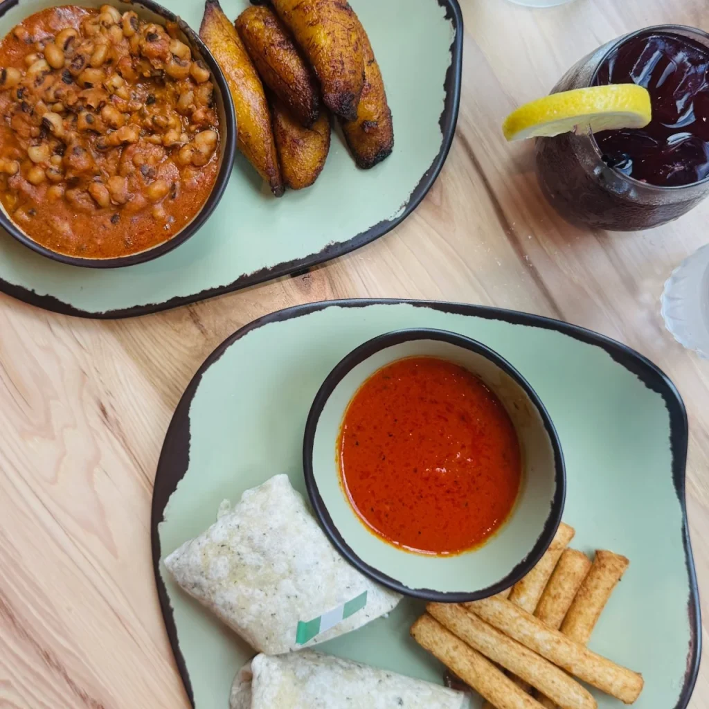 Two plates of food featuring black-eyed peas stew, fried plantains, spicy sauce, and snacks. A drink with lemon sits nearby.