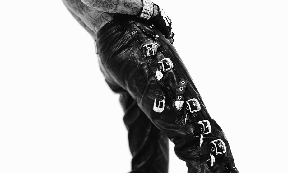 Close-up of a person wearing black leather pants with multiple silver buckles and tattoos visible on their arm.