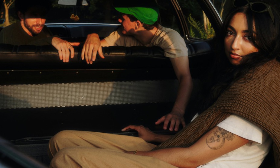 Three people sit in a car, one woman gazes at the camera while two men engage in conversation.
