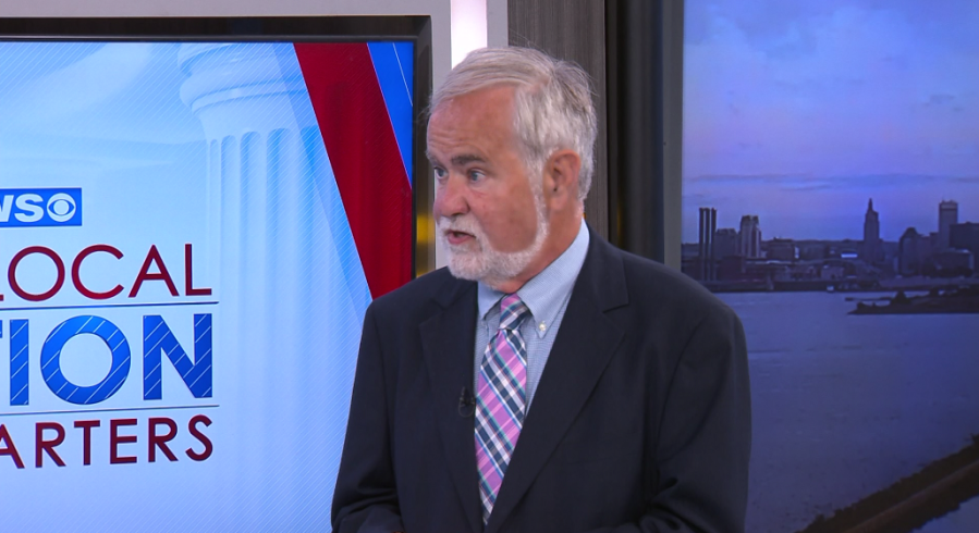 A man with a beard speaks in a studio, with a city skyline visible in the background.