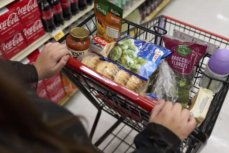 A shopping cart filled with groceries, including pasta, sauce, vegetables, and a drink.