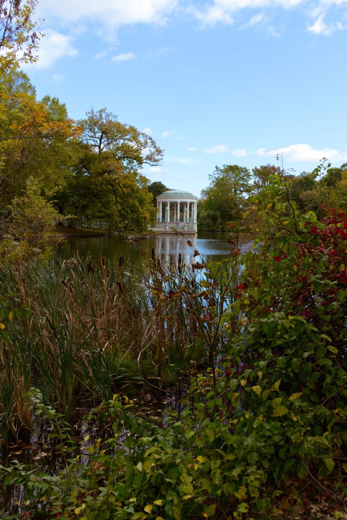 A classical gazebo stands by a tranquil pond, surrounded by colorful foliage and tall grasses.