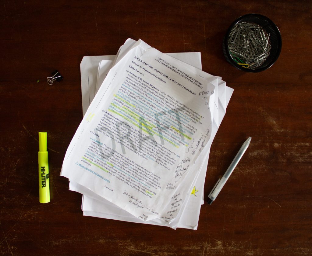 Papers with highlighted text and notes, a highlighter, and a pen on a wooden table. A bowl contains paperclips.