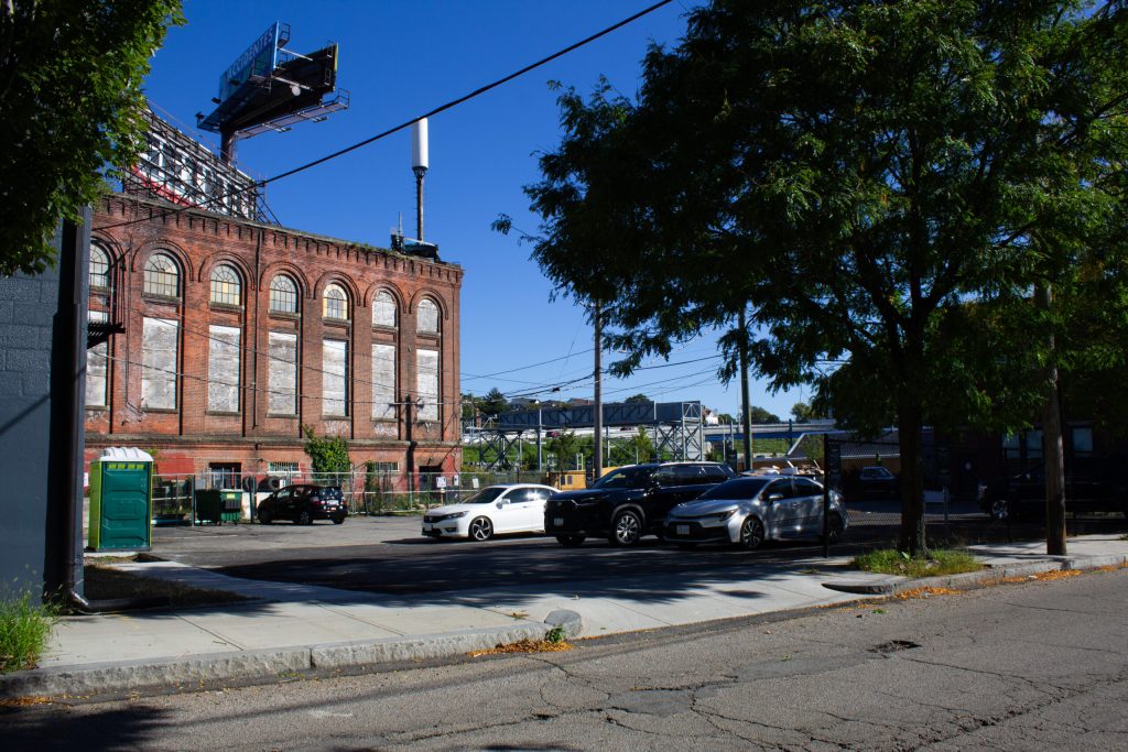 Red brick building with boarded windows, parked cars, and a green portable restroom in a sunny urban setting.