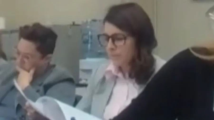 Two people sit at a table, one reading documents while another appears to listen attentively.