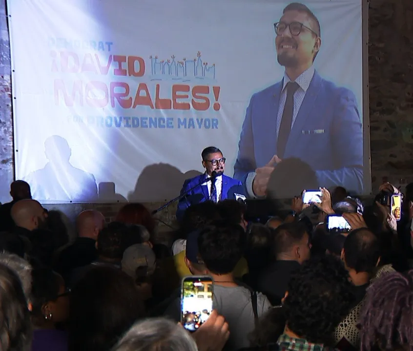 David Morales speaks at a campaign event, with supporters holding phones and a large banner behind him.