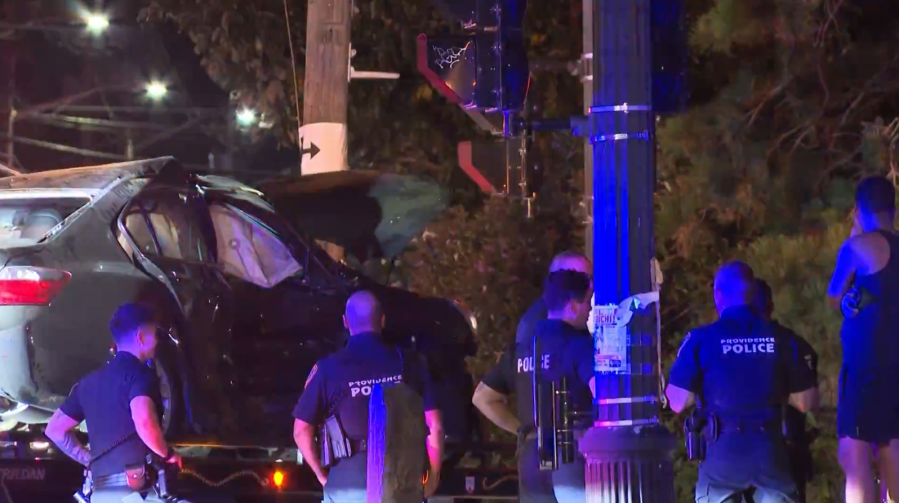 Police officers investigate a damaged vehicle on a tow truck at night, with traffic lights in the background.