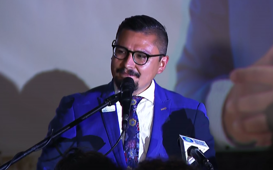 A man in a blue suit speaks into a microphone at an event, with a large screen behind him.