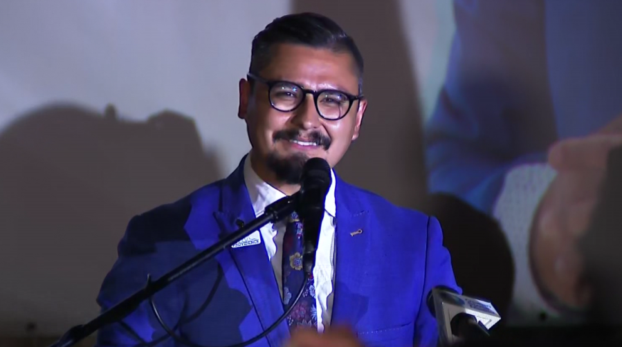 A man in a blue suit smiles while speaking into a microphone at an event.