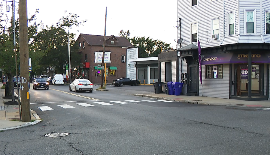 Cars drive on a street with a crosswalk; buildings and trees line the road. A store displays a sign.