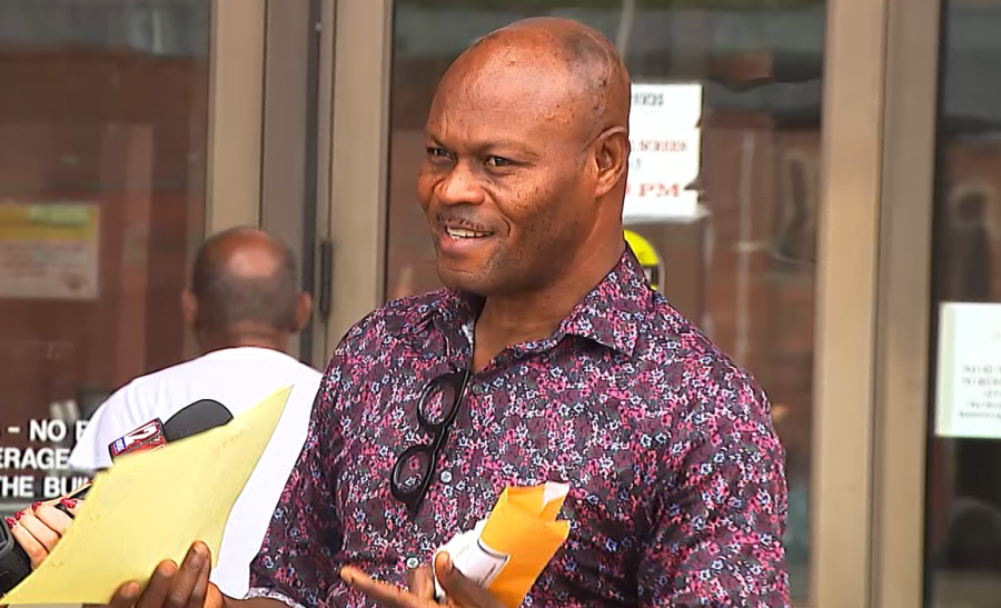 A man in a patterned shirt smiles while holding papers outside a building.
