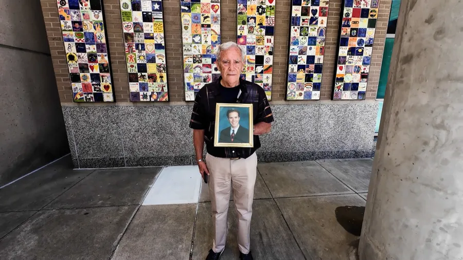 An elderly man holds a framed portrait, standing in front of a colorful tiled wall.