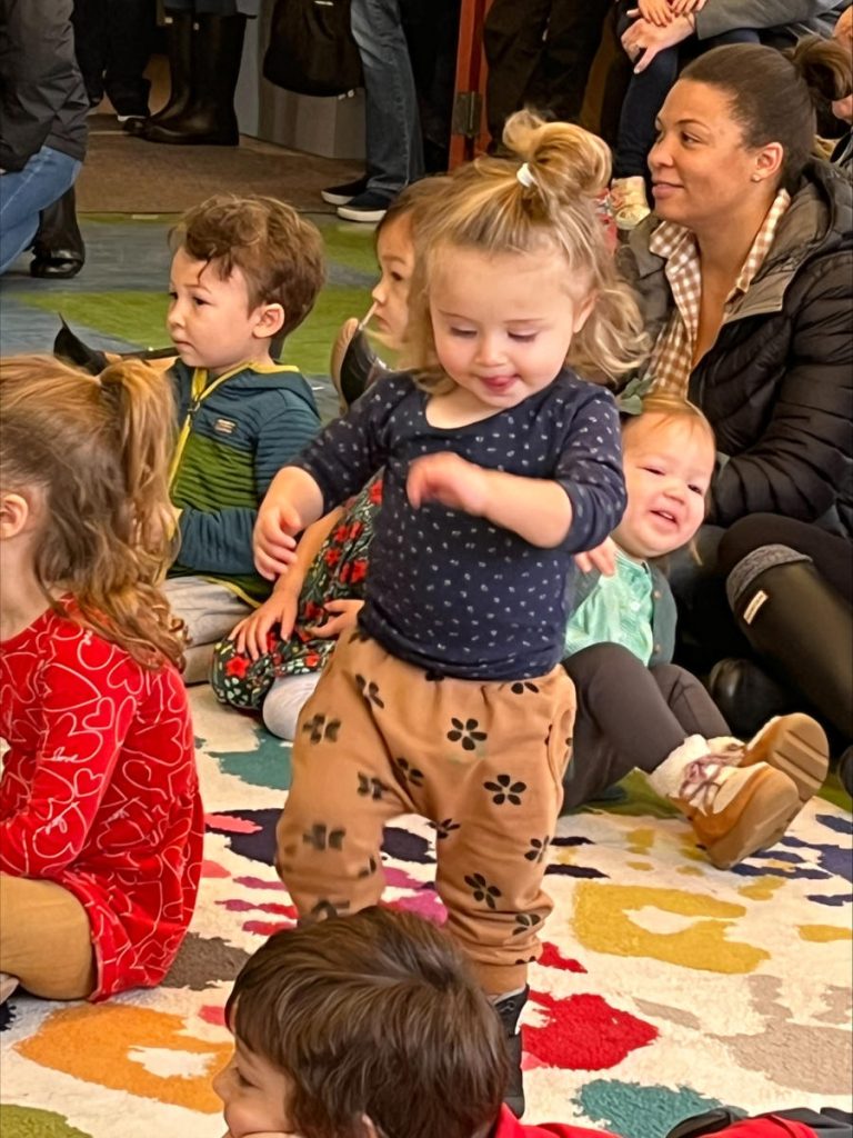 A group of children sit on a colorful rug while one girl dances joyfully in the foreground.