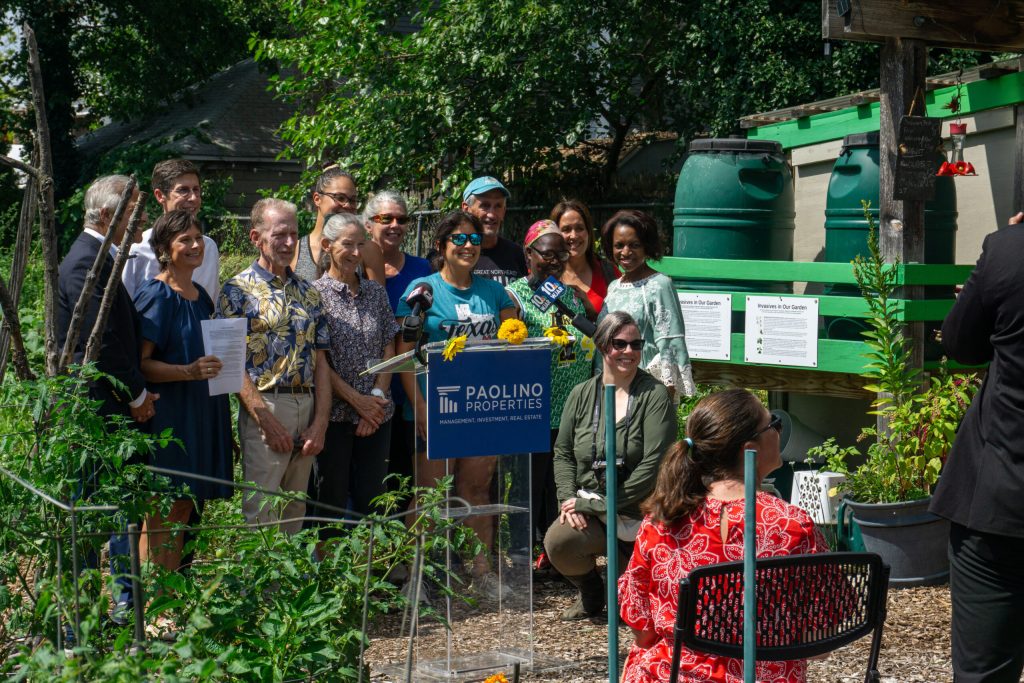 A diverse group of people gathers in a garden, smiling and posing near a podium and informational signs.